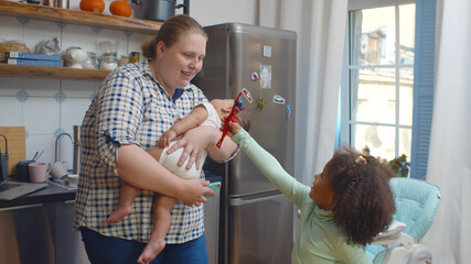 Caucasian mother holding mixed race newborn baby and playing with toddler girl in kitchen