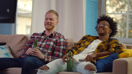 Cheerful multiethnic male friends with beer watching tv at home