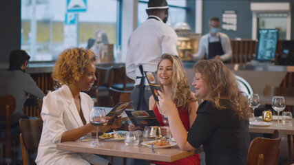 African waiter in safety mask and shield brining menu to young women in restaurant