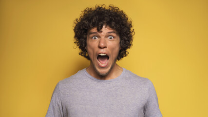 Obraz na płótnie Canvas Emotional young man screams. Caucasian guy opens mouth bulging eyes while looking at camera. Isolated on yellow background.