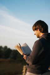 Young man reading a book in the sunlight with trees at the back and clear skies