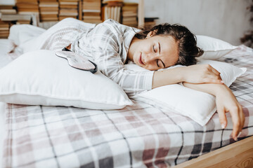Fototapeta na wymiar A sleepy brunette girl lies in bed with a blindfold waking up in the morning. Cozy home morning in cute pajamas. Have a good time, relax and dream. Healthy sleep.