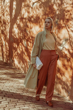 Outdoor, Street Fashion Portrait Of Elegant Woman Wearing Stylish Long Pastel Light Green Trench Coat, Turtleneck, Orange Trousers, Loafers,  Holding Trendy Leather White Padded Bag