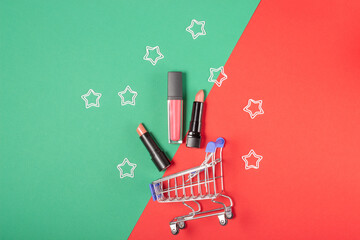 Women's lipsticks and lip glosses in a shopping trolley on a bright red and green background. The concept of buying cosmetics, online shopping, holiday