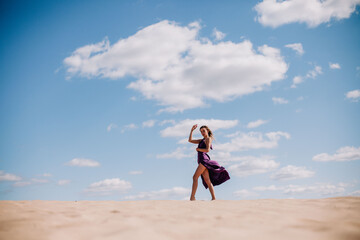 A young, slender girl in a beige dress with purple cloth in her hands posing in the desert in the...