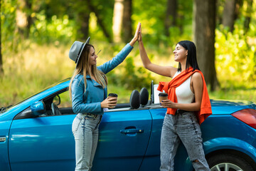 Beautiful young women giving high five, having fun together, cheering with raised arms driving modern convertible car on summer day