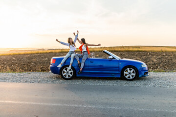 Couple of best women friends having fun together smiling while sitting on convertible car on sunset