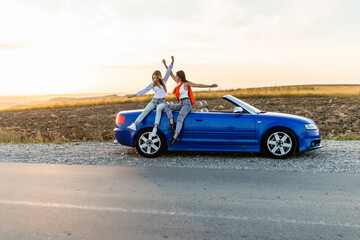 Obraz na płótnie Canvas Couple of best women friends having fun together smiling while sitting on convertible car on sunset