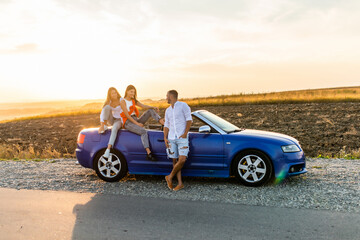 Group of friends having fun in convertible car during road trip at sunset. Young travel people driving a cabriolet during summer holidays. Happiness, vacation and youth lifestyle concept