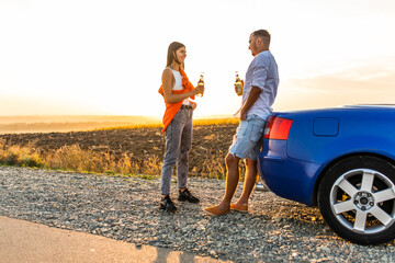 Young pretty couple drinking beer while sitting near convertible and enjoy sunset together