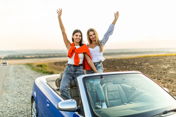 Happy two women in car driving on road trip. Young two women having fun dancing and cheering in car driving on travel vacation together.