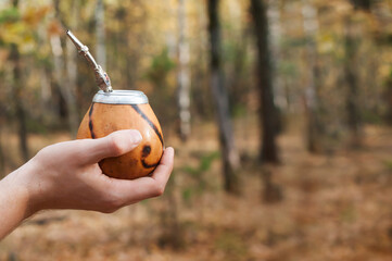 Male hand holds traditional drink yerba mate. Outdoors in the autumn forest. Close-up. Copy space