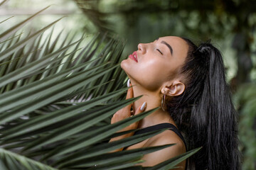 Horizontal beauty shot of pretty sensual Asian girl model, with long black ponytail hair, tounching her neck with eyes closed while posing between exotic palm leaves. Natural beauty and people concept