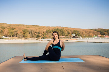 Fototapeta na wymiar a female yogi performs the Marichiasana asana on a blue Mat on a pier with a lake background