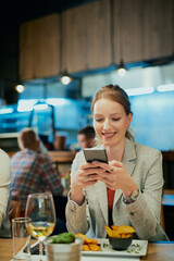 Beautiful smiling woman sitting in restaurant, having dinner and using phone for texting to friends.