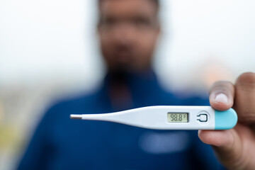 Sick man holds digital thermometer with body temperature reading in Fahrenheit up close to the...