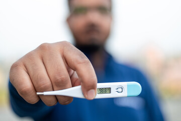 Sick man holds digital thermometer with body temperature reading in Fahrenheit up close to the camera.