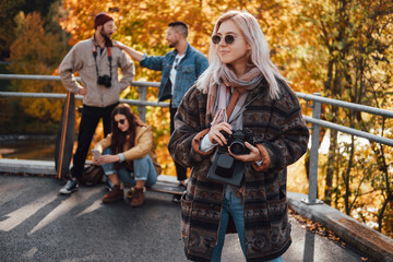 Obraz premium Pretty girl with blond hairs and camera posing in foreground in autumn landscape background and her friends in daytime.