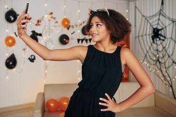 Portrait of pretty smiling teenage girl with cat ears taking selfie at Halloween party