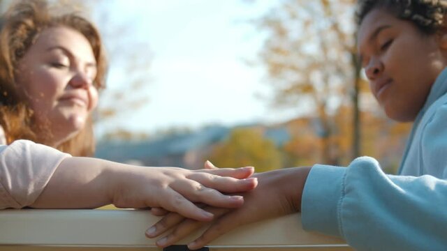 Side view of diverse lesbian couple sitting on bench and holding hands outdoors