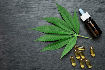Fototapeta na wymiar Capsules or pills and bottle with CBD cannabis or hemp oil on the leaf of marijuana on the black wooden background with copy space. Alternative medicine concept. Cosmetics and skin care products