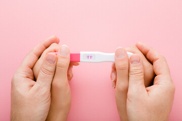 Young adult woman and man hands holding together pregnancy test with two stripes on light pink...