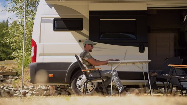Caucasian Men in His 40s Working on His Laptop Computer Outside His RV Camper Van on Summer Camping