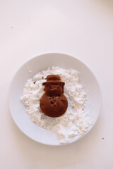 Chocolate cookies in the form of a snowman lie in coconut shavings in a white plate on a white background.