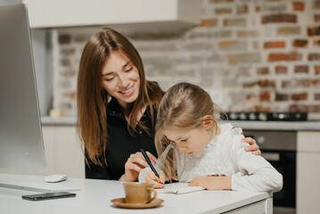 A young smiling mom is hugging a pretty daughter while she is writing in the notebook at the workplace. A gorgeous mother is looking at her blonde child which is doing homework at home.