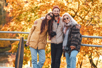 Joufyl company of bearded guy and two beautiful girls posing on bridge in background of autumn forest in daytime.
