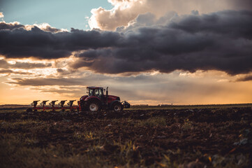 Tractor plowing the fields in the countryside by sunset. Agricultural tractor plowing the field. Red Tractor with plow. Sunset over the autumn field.