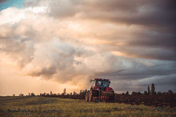 Obraz premium Tractor plowing the fields in the countryside by sunset. Agricultural tractor plowing the field. Red Tractor with plow. Sunset over the autumn field.