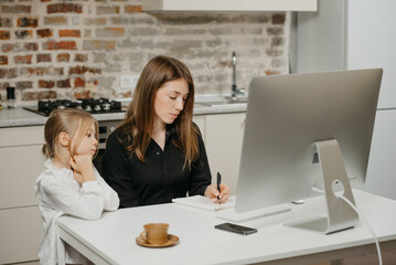 A mother is working remotely at home while a daughter is watching it. A businesswoman is doing notes while working from an apartment near her blonde child.
