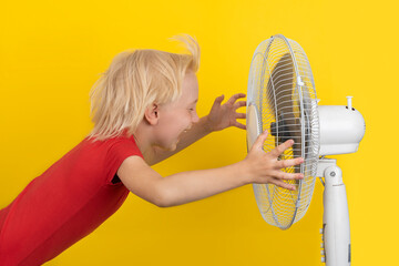 Boy is enjoying the cold air of the fan. Summer heat. Child and fan on yellow background - obrazy, fototapety, plakaty