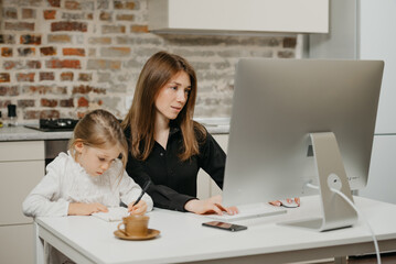 A young mom is working remotely carefully while a daughter is drawing in the notebook at home. A pretty mother is studying on a computer near her blonde child which is writing diligently.