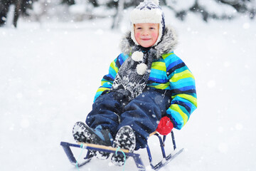 a small child boy in a bright striped jacket sitting on a sledge smiles against the background of snowfall. Winter entertainment