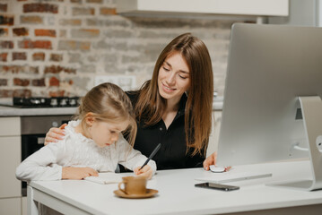 A young mom is hugging a daughter while she is drawing in the notebook at home. A gorgeous mother is looking at her blonde child which is doing homework.