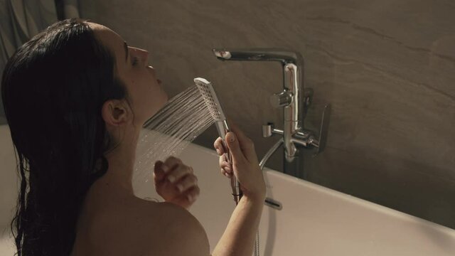 Hot woman sitting under shower streams in bath. Cute girl relaxing in bathtub.