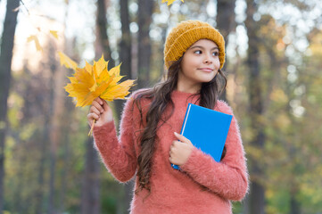 Happy small kid hold school book collecting fall leaves in autumn park outdoors, education