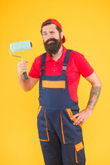 brutal bearded man painter worker in boiler suit uniform using paint roller tool for painting wall into yellow color, creativity