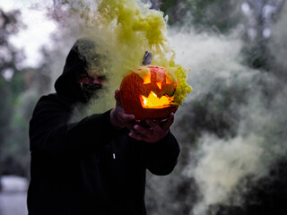 Hombre encapuchado con mascarilla sujetando una calabaza con humo de colores 