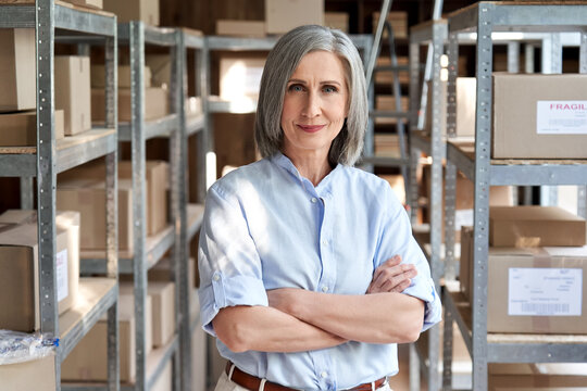 Confident mature older woman retail seller, entrepreneur, online store dropshipping small business owner looking at camera standing in delivery shipping warehouse with parcel boxes, headshot portrait.