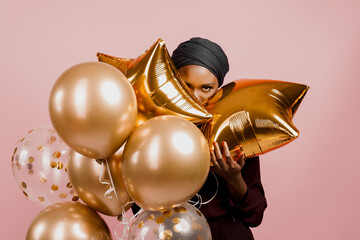 Black girl with golden ballons have a party and smile isolated on pink background. African woman celebrate graduation. Happy emotions of muslim young woman