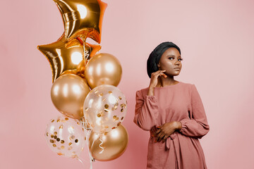 Christmas celebration. African american holiday. Attractive girl with golden helium balloons isolated peach background. Young black woman weared traditional dress and scarf.