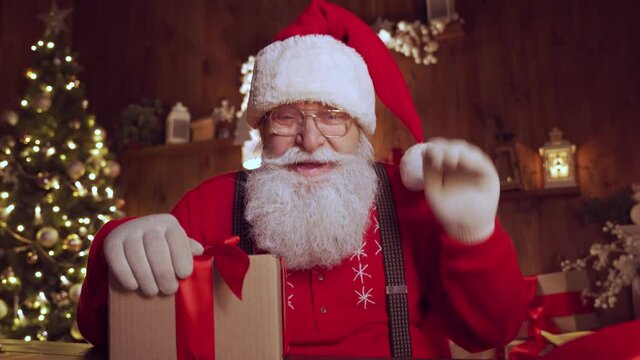 Happy Santa Claus, Saint Nicholas waving hand holding present talking to camera video calling kid or recording Merry Christmas virtual greetings on xmas eve sitting at table at home, webcam view.