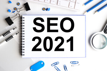 seo 2021, text on white notepad near keyboard, blue pencils, black magnifier. on white background. top view