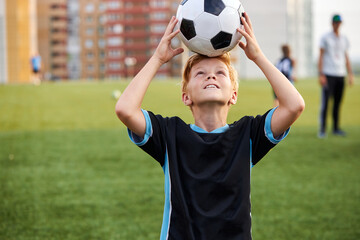 portrait of teen boy having fun with ball in the stadium, he holds soccer ball on head, take a break before game