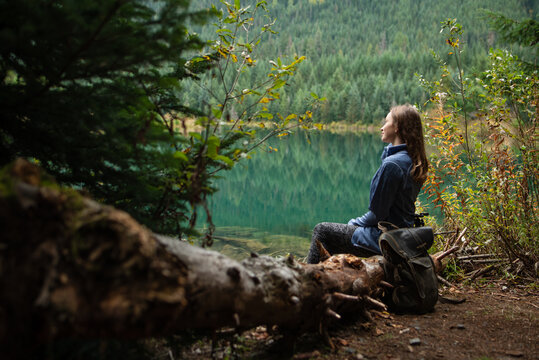 Young travel women siting on deadwood near amazing lake on coniferous forest background. Concept of mountains trip. Side view. Meditation in nature. Relax. Travel journal. Fantastic weekends.