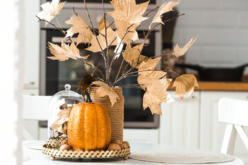 Branches with Golden leaves and a pumpkin on a tray. In the background-the interior of a white kitchen in the Scandinavian style. The concept of home comfort. Autumn decor for the Halloween holiday.