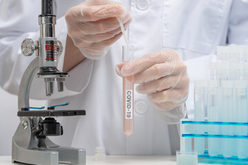 Close-up view of scientist in protection gloves pours vaccine in biological tube with label covid-19 on laboratory room background. Healthcare and medical concept.